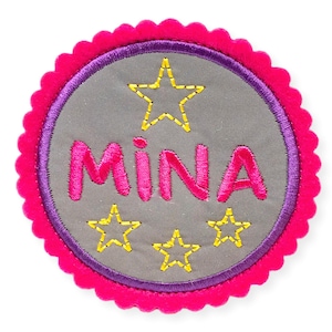 Könnte beinhalten: Ein rosa-violetter Filz-Kreis mit einem gewellten Rand. Die Mitte des Kreises hat einen grauen Hintergrund mit dem Namen "MINA", der in rosa gestickt ist, und vier gelben Sternen.