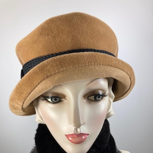 May include: A tan cloche hat with a black band and a black faux fur scarf. The hat has a rounded crown and a small brim. The scarf is dark brown and fluffy. The items are displayed on a mannequin head.