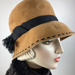 May include: A tan cloche hat with a black woven band and tassel. The hat features a stitched brim detail and is displayed on a mannequin head. The hat is a vintage style.