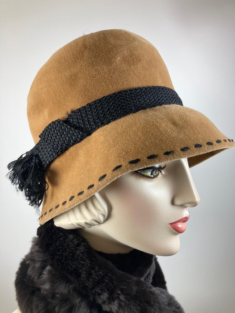 May include: A tan cloche hat with a black woven band and tassel. The hat features a stitched brim detail and is displayed on a mannequin head. The hat is a vintage style.