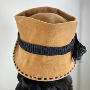 May include: A tan-colored hat with a black band and tassel. The hat has a rounded crown and a wide brim with decorative stitching. The black band encircles the hat, and a black tassel hangs on the side. The hat appears to be made of a soft, suede-like material.