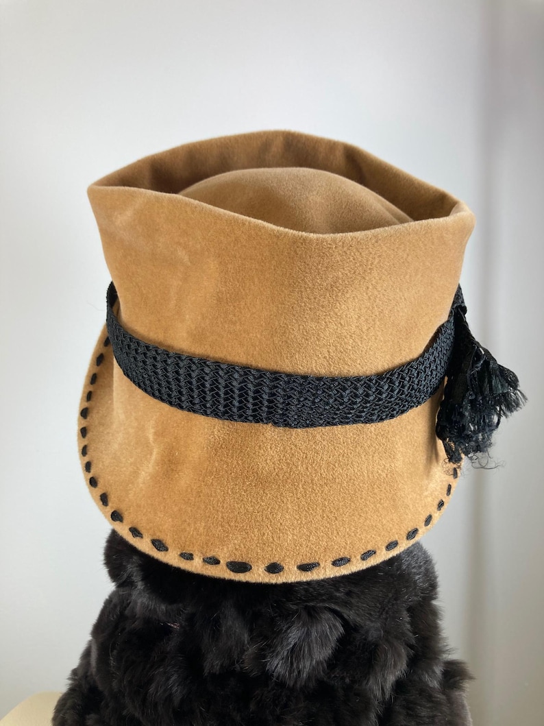 May include: A tan-colored hat with a black band and tassel. The hat has a rounded crown and a wide brim with decorative stitching. The black band encircles the hat, and a black tassel hangs on the side. The hat appears to be made of a soft, suede-like material.