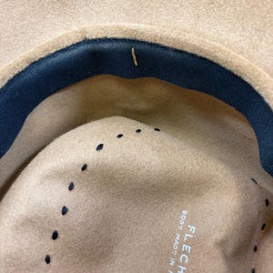 May include: Close-up of the interior of a tan fedora hat with a dark blue band and black stitching. The hat is labeled "FLECHET BODY MADE IN FRANCE".