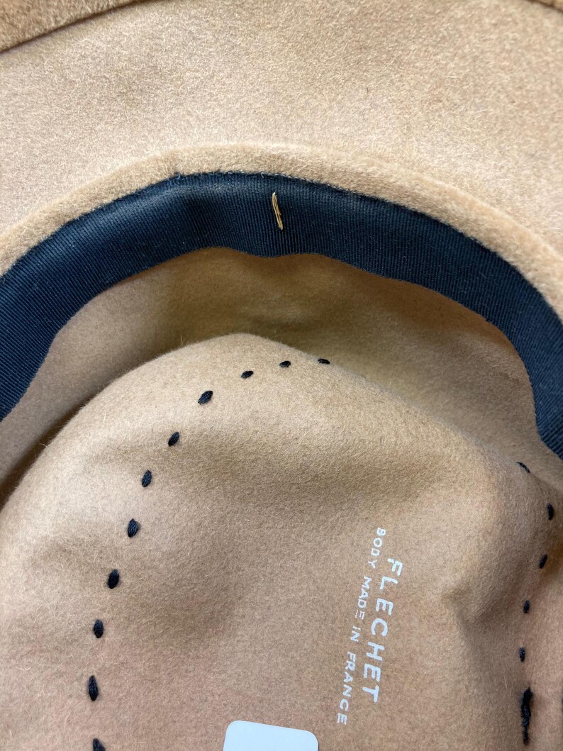 May include: Close-up of the interior of a tan fedora hat with a dark blue band and black stitching. The hat is labeled "FLECHET BODY MADE IN FRANCE".