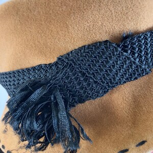 May include: A tan cloche hat with a black woven band and tassel detail. The hat features a stitched brim and a soft, textured appearance. This vintage-style hat is a classic accessory.