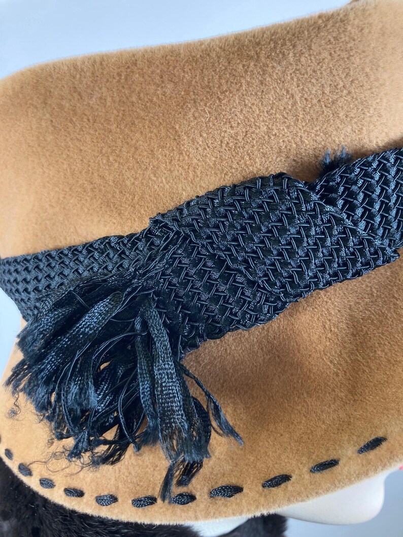 May include: A tan cloche hat with a black woven band and tassel detail. The hat features a stitched brim and a soft, textured appearance. This vintage-style hat is a classic accessory.