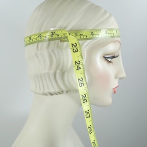 May include: A mannequin head with a cream-colored complexion is measured with a yellow measuring tape. The tape wraps around the head, showing measurements in inches. The tape extends down the side of the head, displaying measurements up to 30 inches.