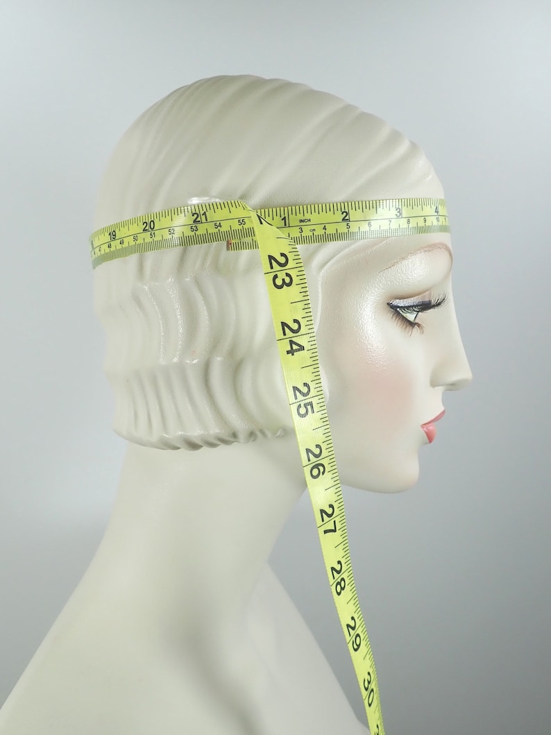 May include: A mannequin head with a cream-colored complexion is measured with a yellow measuring tape. The tape wraps around the head, showing measurements in inches. The tape extends down the side of the head, displaying measurements up to 30 inches.