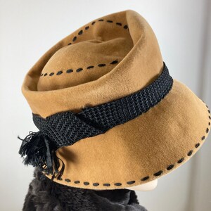 May include: A tan-colored hat with a black woven band and tassel. The hat features black stitching around the brim and crown. The hat is likely vintage or retro in style.