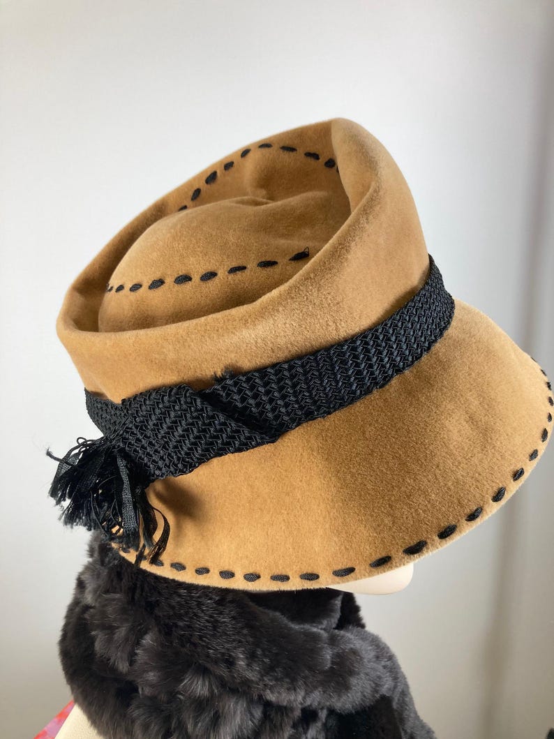 May include: A tan-colored hat with a black woven band and tassel. The hat features black stitching around the brim and crown. The hat is likely vintage or retro in style.
