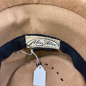 May include: A tan-colored hat with a black ribbon and a tag that reads "Miss Alice". The hat has a classic design with a rounded crown and a wide brim. The hat is made in France.
