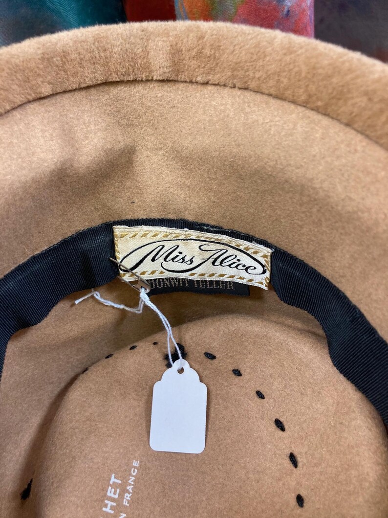 May include: A tan-colored hat with a black ribbon and a tag that reads "Miss Alice". The hat has a classic design with a rounded crown and a wide brim. The hat is made in France.