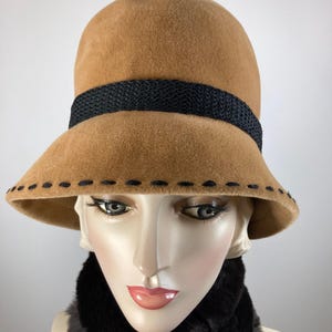 May include: A tan cloche hat with a black woven band and decorative stitching along the brim. The hat is displayed on a mannequin head with a black faux fur scarf.
