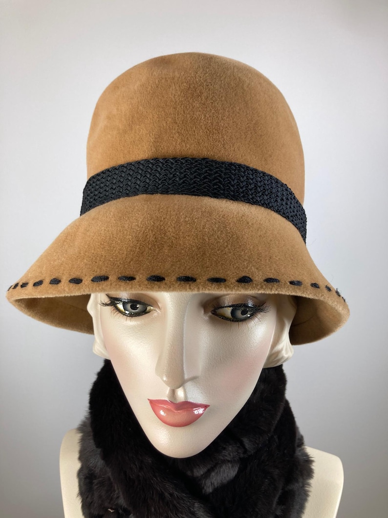 May include: A tan cloche hat with a black woven band and decorative stitching along the brim. The hat is displayed on a mannequin head with a black faux fur scarf.