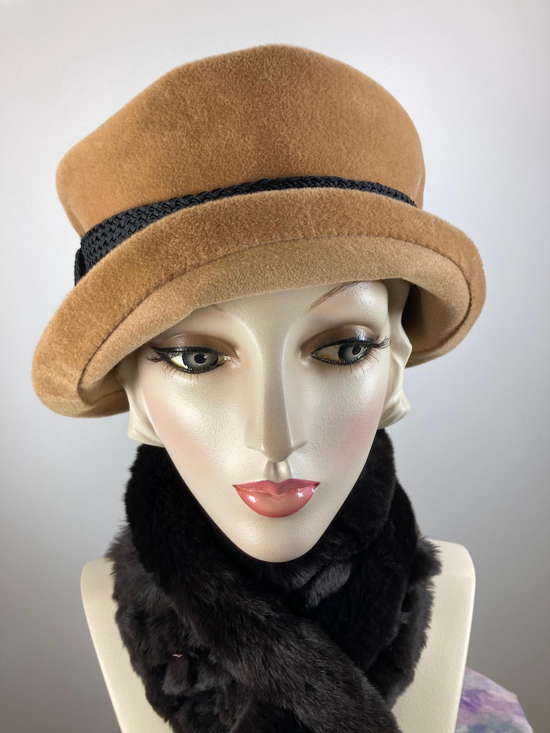 May include: A tan cloche hat with a black band and a black faux fur scarf. The hat has a rounded crown and a small brim. The scarf is dark brown and fluffy. The items are displayed on a mannequin head.