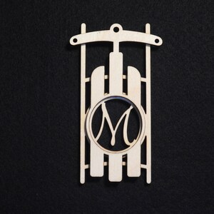 May include: A wooden sled-shaped ornament with the letter "M" in a circular frame. The ornament is made of light-colored wood and has a hole at the top for hanging. The background is black.