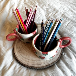 May include: Two ceramic mugs with strawberry patterns, filled with coloured pencils, sit on a wooden slice. The mugs have pink handles and red interiors. The pencils are various colours, including red, yellow, and blue. The background is a white, textured surface.