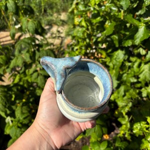 May include: A ceramic mug featuring a speckled off-white interior and a blue-toned exterior. The handle is shaped like a whale tail, extending from the rim. The glaze has a mottled texture with darker blue accents along the rim and the tail handle.
