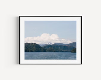 May include: A framed landscape photograph featuring a deep blue body of water in the foreground, with dense green forested hills rising in the distance under a pale blue sky with white clouds and a single small bird visible in the upper left.