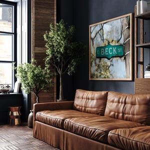 May include: A living room scene featuring a brown leather sofa, a framed picture of a street sign that reads "E BECK ST", and two small green trees. The room has a dark blue wall and a wooden floor.