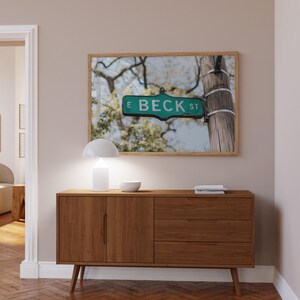 May include: A framed print of a street sign reading "E BECK ST" in green and white, mounted on a wooden post. The print hangs above a mid-century modern wooden cabinet with a white lamp and bowl.