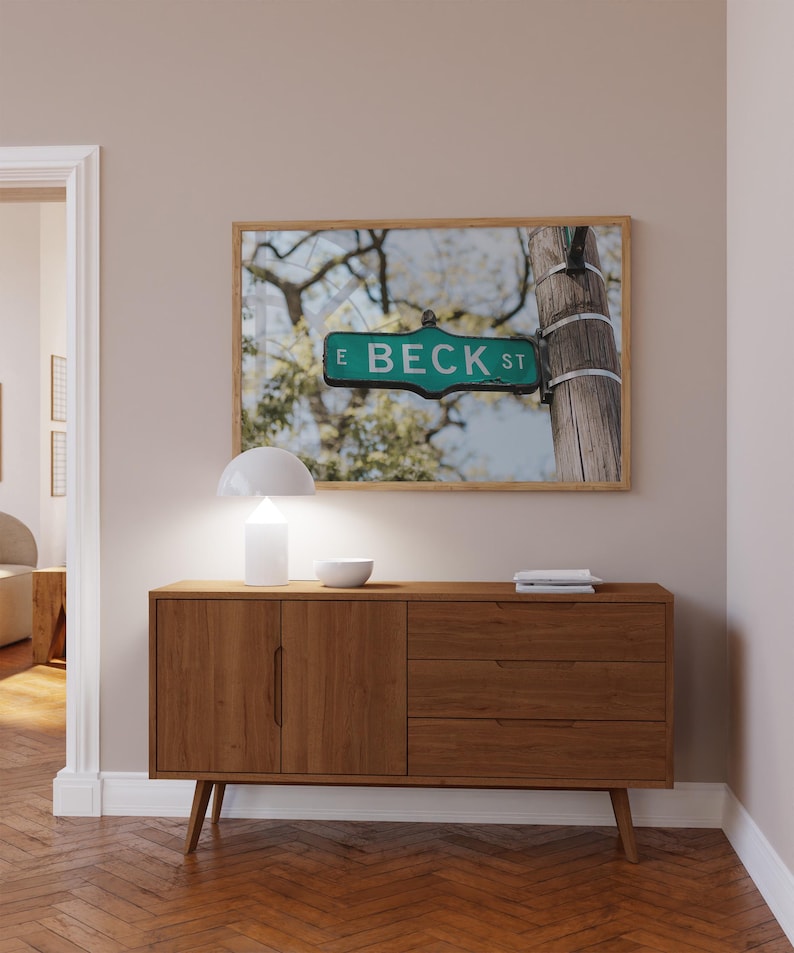 May include: A framed print of a street sign reading "E BECK ST" in green and white, mounted on a wooden post. The print hangs above a mid-century modern wooden cabinet with a white lamp and bowl.