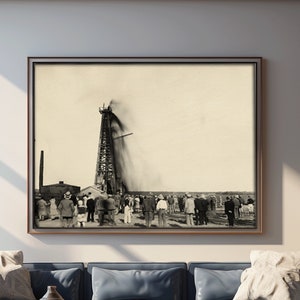 May include: A black and white photograph of a large oil derrick with smoke billowing from the top. A crowd of people stand in front of the derrick, watching the scene unfold. The photograph is framed and hanging on a white wall in a living room setting.