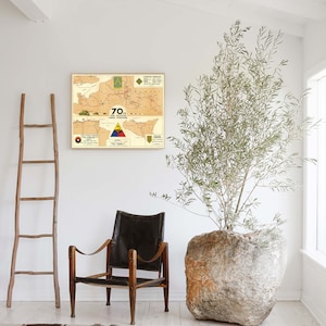 May include: A vintage map print of a military operation, framed in a light wood frame, hangs on a white wall. A brown leather armchair sits in front of the map, and a large, round, light-colored stone planter with a green leafy plant sits to the right of the chair.