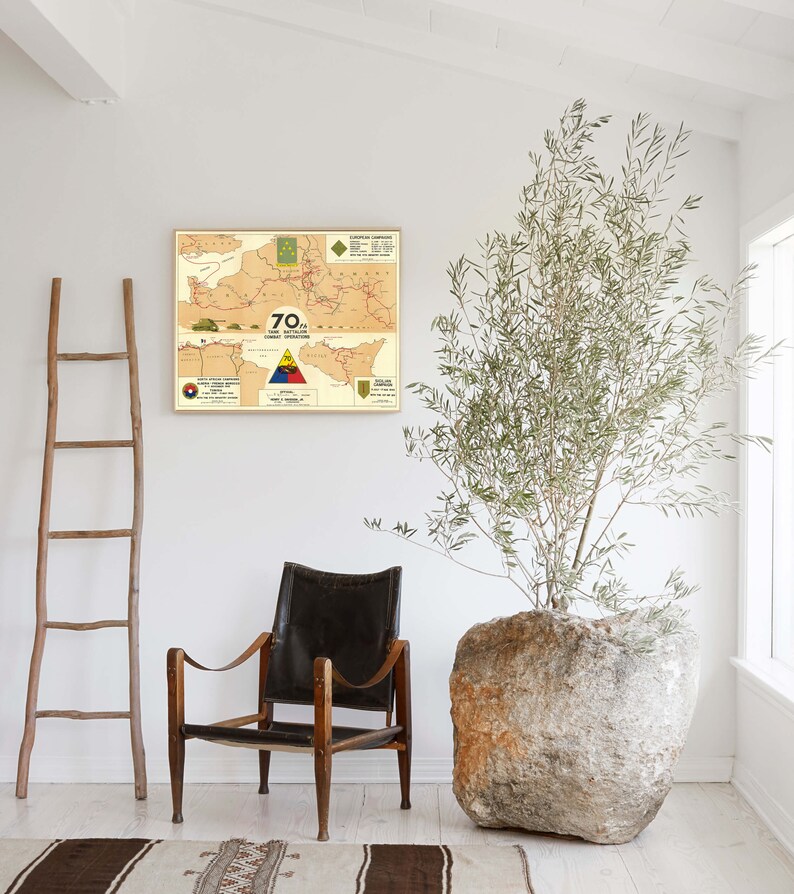 May include: A vintage map print of a military operation, framed in a light wood frame, hangs on a white wall. A brown leather armchair sits in front of the map, and a large, round, light-colored stone planter with a green leafy plant sits to the right of the chair.