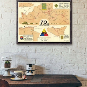 May include: A vintage map showing the combat operations of the 70th Tank Battalion during World War II. The map shows the battalion's movements across North Africa, Sicily, and Europe. The map is framed in a dark wood frame and is hanging on a white brick wall.