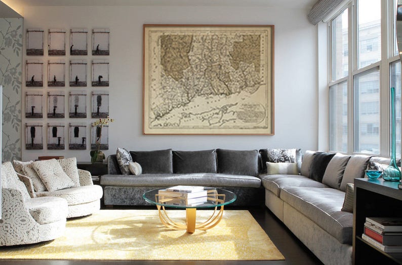 May include: A gray sectional sofa with multiple pillows sits in a living room with a glass coffee table and a framed map of New England on the wall.