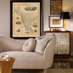 May include: A beige chaise lounge with two pillows, one with a button tuft and the other with a geometric pattern. A gold side table with a crystal object on top sits in front of the chaise. A framed map of South America hangs on the wall above the chaise. A gold-framed picture with a gold-toned image hangs on the wall to the right of the map. A black lamp with a black lampshade sits on a wooden cabinet with glass doors.