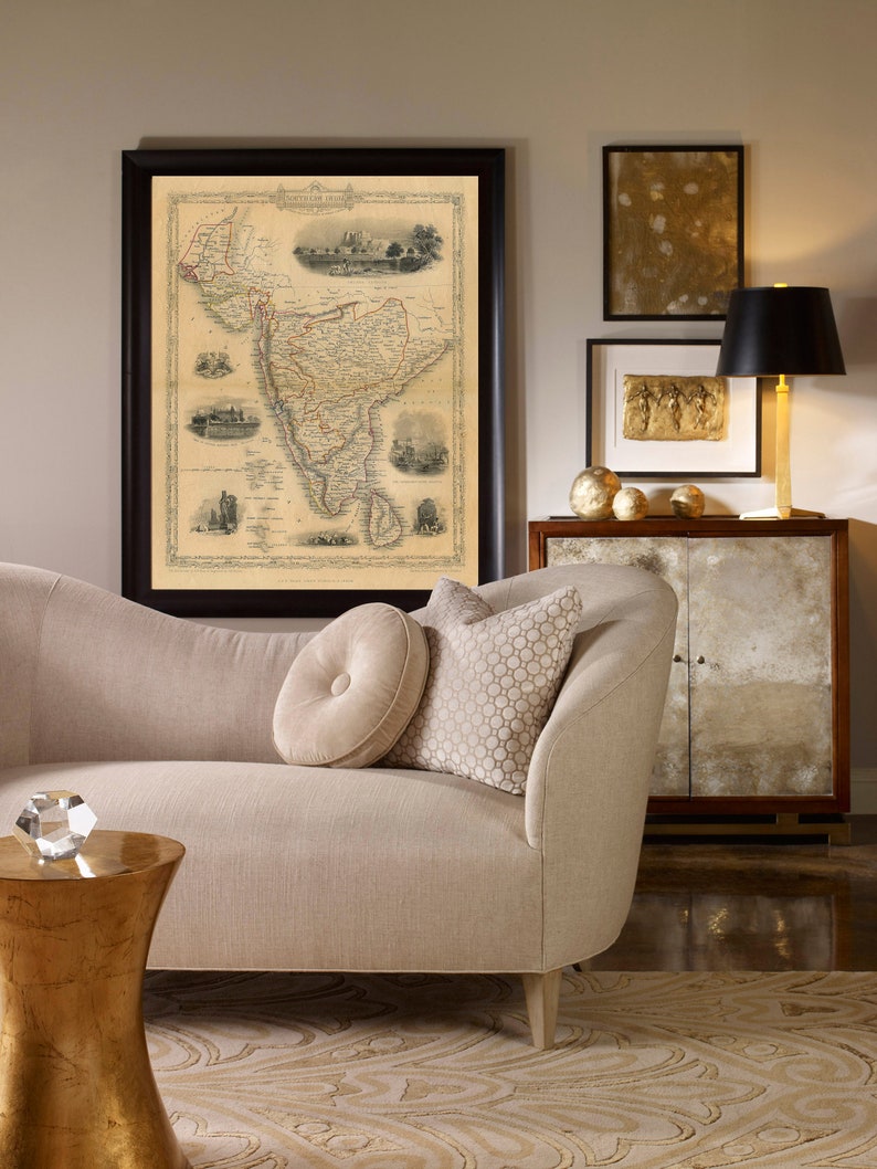 May include: A beige chaise lounge with two pillows, one with a button tuft and the other with a geometric pattern. A gold side table with a crystal object on top sits in front of the chaise. A framed map of South America hangs on the wall above the chaise. A gold-framed picture with a gold-toned image hangs on the wall to the right of the map. A black lamp with a black lampshade sits on a wooden cabinet with glass doors.