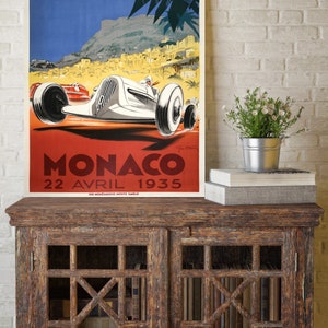 May include: Vintage poster advertising the 7th Grand Prix Automobile de Monaco, held on April 22, 1935. The poster features a white race car speeding through a winding road with a view of the city of Monaco in the background.