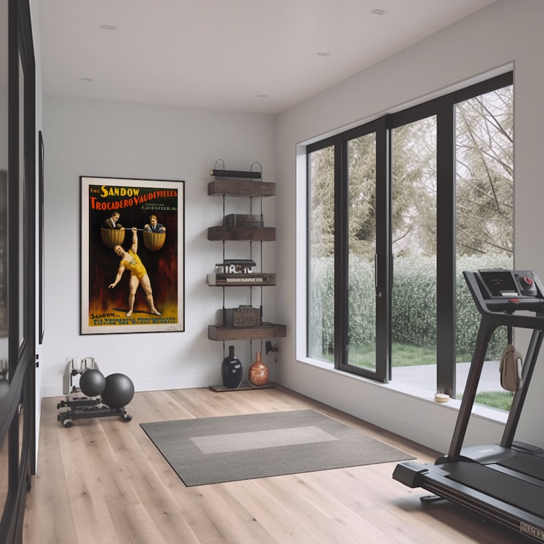 Puede incluir: Un cartel vintage que anuncia "The Sandow Trocadero Vaudevilles" con un hombre fuerte levantando dos hombres en cubos. El cartel est&aacute; enmarcado y colgado en una pared blanca en un gimnasio en casa con una cinta de correr y otros equipos de ejercicio.