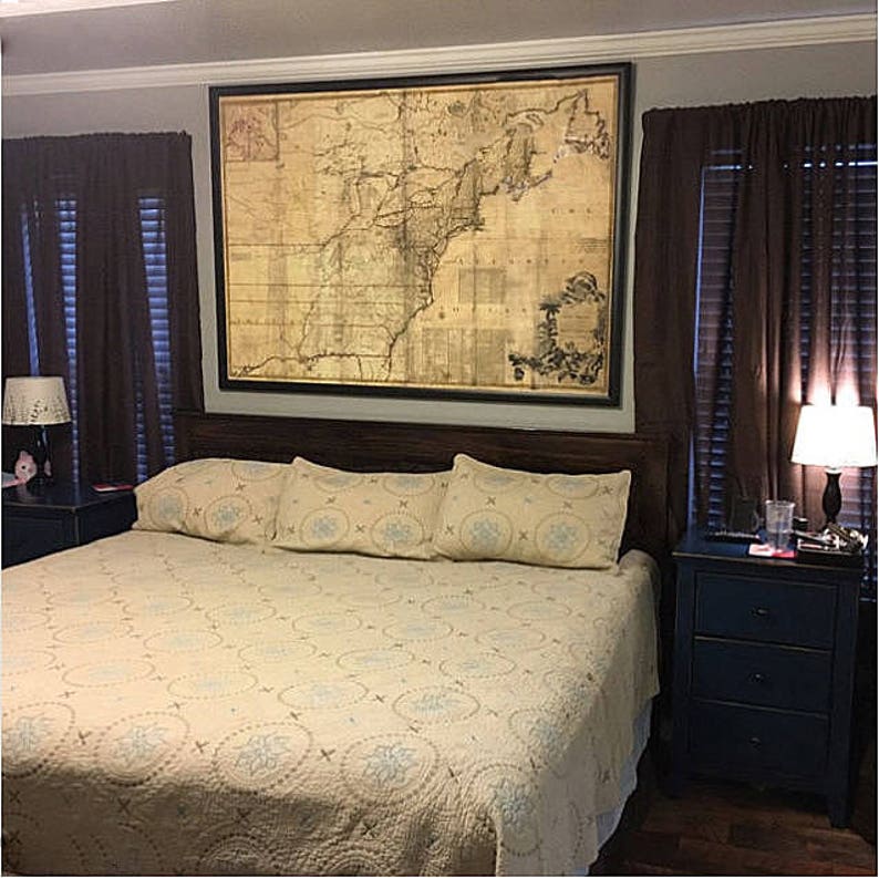 May include: A bedroom with a large framed map of the United States hanging above the headboard of a bed with a beige quilt and blue and white patterned pillows. There are two nightstands with lamps on either side of the bed.