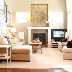 May include: A well-lit living room with a neutral color palette. Features include a beige sectional sofa, a fireplace with a framed map above, a large clock, and a TV. The room is furnished with lamps, a coffee table, and two armchairs.