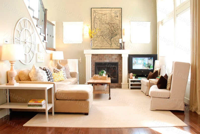May include: A well-lit living room with a neutral color palette. Features include a beige sectional sofa, a fireplace with a framed map above, a large clock, and a TV. The room is furnished with lamps, a coffee table, and two armchairs.