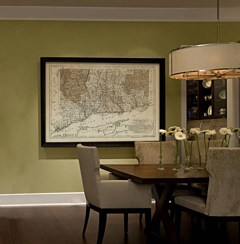 May include: A dining room with a large framed map of Connecticut hanging on a green wall. The map is in black and white and shows the state's borders and major cities. A wooden table with six chairs is in the center of the room. The table is set with white plates and silverware. There are white flowers in vases on the table.