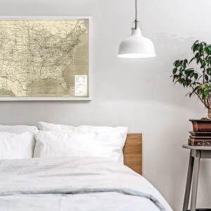 Puede incluir: Un mapa de los Estados Unidos enmarcado en blanco con una luz colgante blanca sobre una cama blanca con una cabecera de madera. Una peque&ntilde;a planta en maceta se encuentra en un taburete gris a la derecha de la cama.