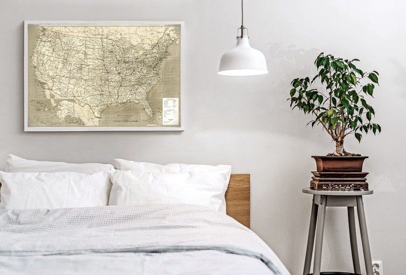 Puede incluir: Un mapa de los Estados Unidos enmarcado en blanco con una luz colgante blanca sobre una cama blanca con una cabecera de madera. Una peque&ntilde;a planta en maceta se encuentra en un taburete gris a la derecha de la cama.