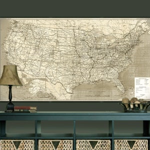 Puede incluir: Un mapa vintage de los Estados Unidos que muestra las l&iacute;neas de ferrocarril y las principales ciudades. El mapa est&aacute; impreso en papel de color crema y enmarcado en un marco negro sencillo. El mapa est&aacute; colgado en una pared verde oscuro sobre una consola azul oscuro con cestas de mimbre.