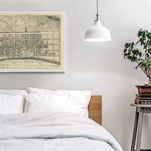 May include: A vintage black and white map of New Orleans, Louisiana, with the title "SITE & LOTS of NEW ORLEANS, PLAN of NEW ORLEANS the CAPITAL of LOUISIANA." The map is framed and hanging on a white wall above a white bed with a wooden headboard. A small potted plant sits on a gray side table.