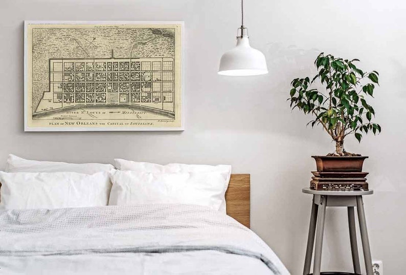 May include: A vintage black and white map of New Orleans, Louisiana, with the title "SITE & LOTS of NEW ORLEANS, PLAN of NEW ORLEANS the CAPITAL of LOUISIANA." The map is framed and hanging on a white wall above a white bed with a wooden headboard. A small potted plant sits on a gray side table.