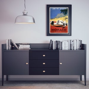 May include: A black framed poster with a vintage design advertising the 7th Grand Prix Automobile Monaco on April 22, 1935. The poster features a white race car with red accents driving on a red road with a mountain and palm trees in the background.