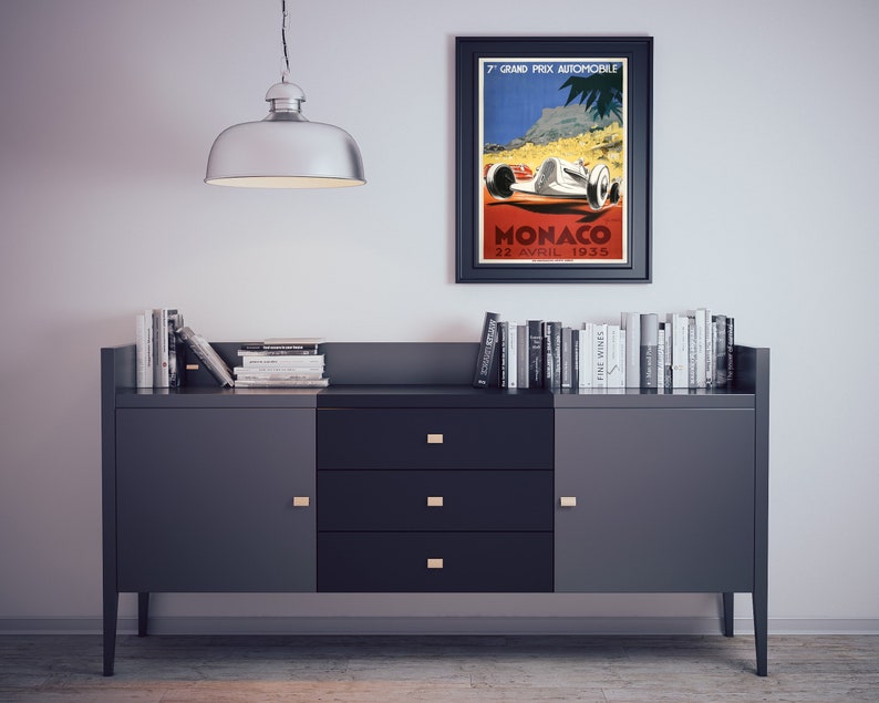 May include: A black framed poster with a vintage design advertising the 7th Grand Prix Automobile Monaco on April 22, 1935. The poster features a white race car with red accents driving on a red road with a mountain and palm trees in the background.