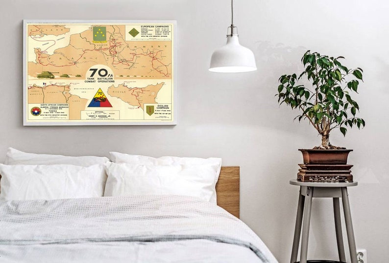 May include: A vintage map showing the combat operations of the 70th Tank Battalion during World War II. The map shows the battalion's movements across Europe and Sicily, with key locations and dates marked. The map is framed and displayed on a white wall.