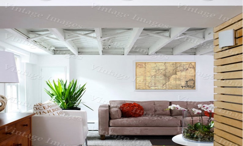 May include: A living room with a white exposed beam ceiling, a brown sofa with a red throw pillow, a large framed map on the wall, a potted plant, and a table with a vase of white orchids.