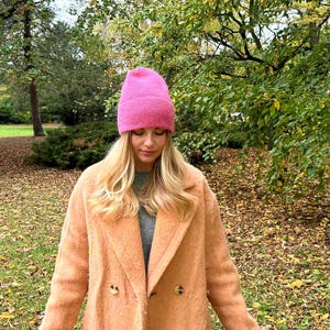 May include: A woman wearing a pink knit beanie and a peach-colored double-breasted coat. The coat has large buttons and a classic lapel. The background features trees with green and yellow leaves, suggesting an autumn setting.