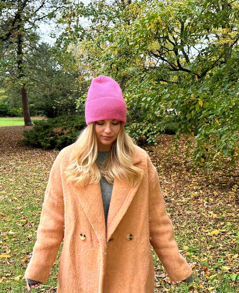 May include: A woman wearing a pink knit beanie and a peach-colored double-breasted coat. The coat has large buttons and a classic lapel. The background features trees with green and yellow leaves, suggesting an autumn setting.